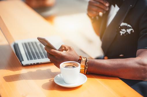 Close-up view of the hand of a black man entrepreneur in a formal summer costume using a smartphone in a street cafe, with a cup of coffee and a laptop next to him on the table on a warm sunny day