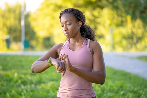 African american fit woman checking fitness tracker, training in urban park, medium shot. Sportswoman looking at smart watch on wrist, jogging outdoors, front view