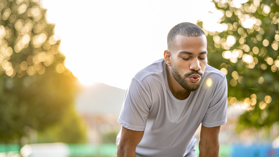 Young african american sportive man bending, breathing deeply after outddor training, close up. Tired person standing, jogging in park, medium shot