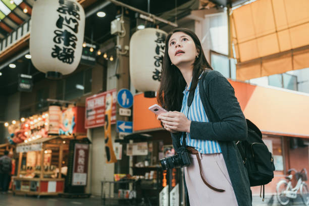 istock lost asian girl using navigation app on cellphone and looking up for information signs while visiting kuromon ichiba market in Osaka japan. translation:â kuromon ichiba marketâ 1436868153