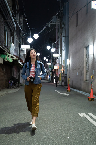full length with vertical view leisure asian girl photographer looking around cityscape while taking walk on shopping street in Osaka, japan at a cool evening in spring