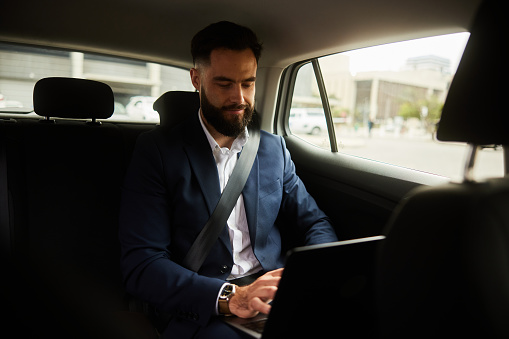 Young businessman sitting in the backseat of a taxi and working on a laptop while going to an appointment