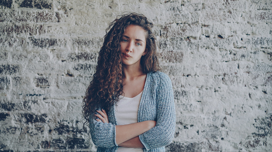 Portrait of angry young lady looking at camera, frowning and expressing disappointment, anger and disapproval standing with arms crossed with nice brick wall in background.