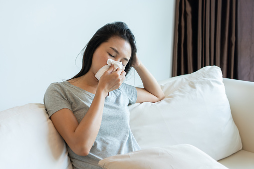 Joven asiática enferma sentada en el sofá sonándose la nariz en un pañuelo conceptual de atención médica, gripe estacional, rinitis alérgica o reacción alérgica en la fiebre del heno. Primer plano, espacio de copia photo