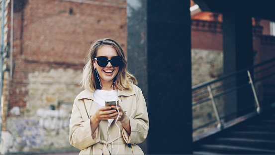 Pretty girl is listening to music through earphones using smartphone and dancing walking in the street and enjoying rhythm and modern city. Girl is wearing sunglasses and trendy coat.