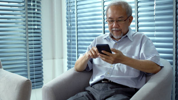 istock Elderly asian man touching screen mobile phone search web or social online sitting on sofa at home. Grandfather male holding cellphone watching something sitting on couch in room. Lifestyle technology 1436892294
