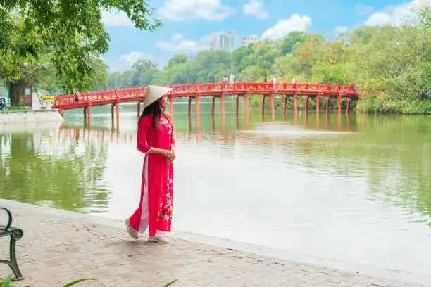 Young Asian woman tourist wearing Ao Dai (traditional Vietnamese dress) sightseeing at the bridge Red Bridge in Hoan Kiem Lake, Hanoi, Vietnam. Copy space