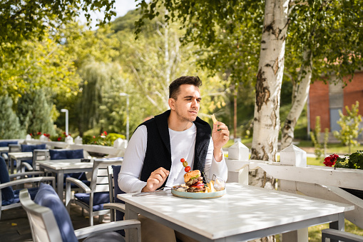 One man young adult caucasian male sitting at the table at restaurant outdoor terrace with burgher and french fires potato chips eat alone in day real people copy space
