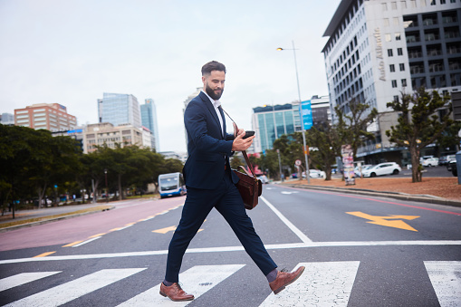 Smiling young businessman checking his phone and a carrying a satchel crossing a street in the city