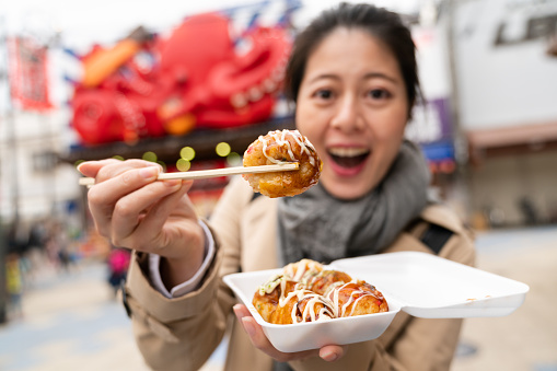 selective focus of delicious takoyaki octopus ball held on chopsticks by an amazed Asian Japanese woman and shown to camera in Shinsekai area in Osaka Japan