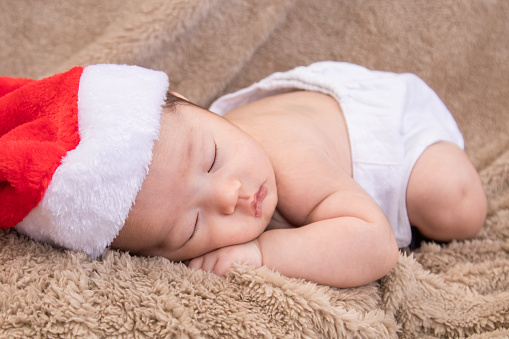 Asian newborn baby wearing christmas hat sleeping on brown blanket, adorable infant lying on bed at home with trust and safe. Little child was born on the same day as Jesus.