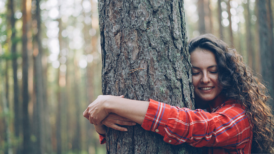 Beautiful young woman with curly hair wearing bright shirt is hugging tree enjoying nature and smiling with closed eyes. Joyful people, recreation and happiness concept.