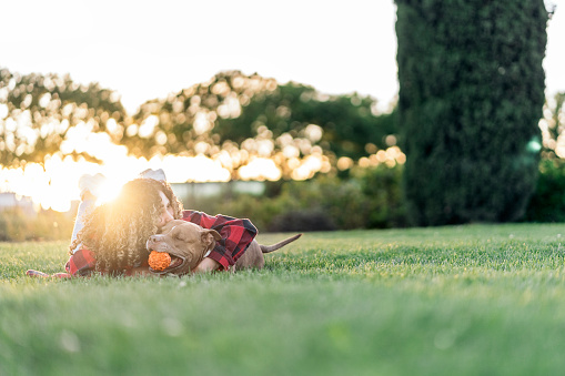 Young african woman with curly hair lying down in the park and kissing her purebred dog.