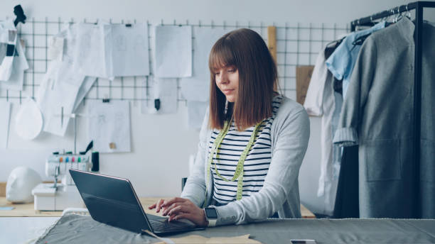 istock Young self-employed tailor is communicating with customers using laptop. Good view of light modern studio in background. Successful e-commerce in clothing design industry concept. 1436904638