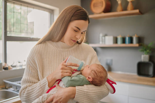 Newborn, baby and mother milk bottle drink of a young mama in a home kitchen with love and care. Calm happiness of a mom holding her child in a family house while her kid is drinking formula stock photo
