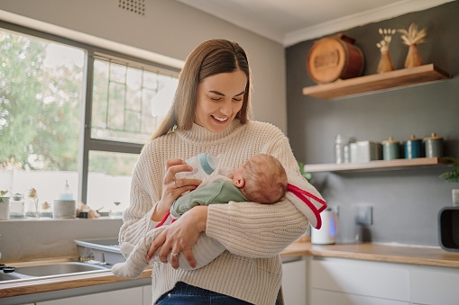 Mother feeding her baby a bottle of milk with love, care and happiness while standing in the kitchen. Happy, smile and newborn child drinking infant formula from his mom in their modern family home.