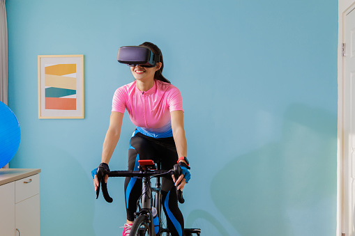 woman cycling with VR glasses in her room - asian female cyclist is playing online bike games