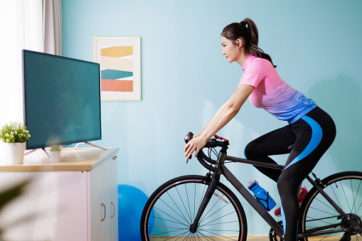 asian female cyclist is exercising in the house by cycling on trainer and playing online bike games with TV