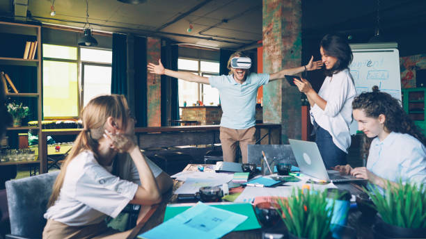 istock Young man is using virtual reality glasses in modern office, his colleagues are working with tablet and laptop, looking at him and laughing. Modern technology and fun concept. 1436907318