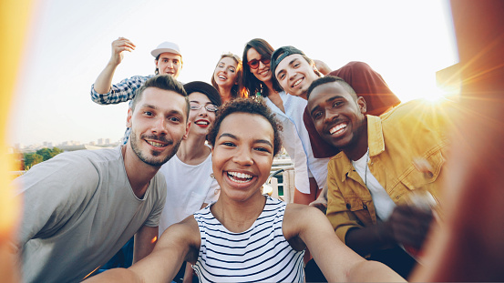 Point of view shot of pretty African American girl holding camera and taking selfie with happy friends at party on roof. Men and women are looking at camera, posing and laughing.