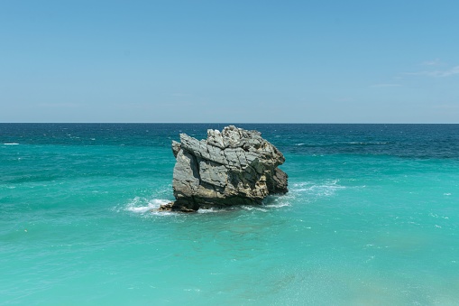 A seascape view of a huge rock in the middle of turquoise sea under clear blue sky