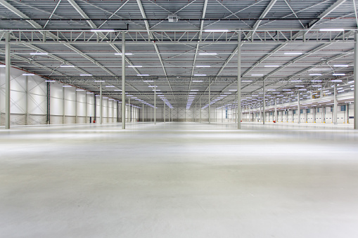 Side view of an empty large warehouse interior with white brick side walls and rough floor illuminated by natural light from ceiling lights and large roof windows. Focus on the side walls. 3D rendered image.