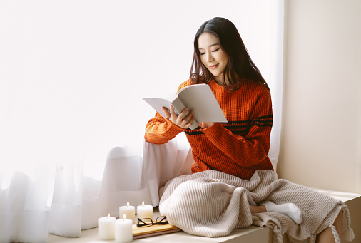 Happy beautiful Asian woman wearing knitted sweater relaxing reading book sitting on windowsill in room decorated for celebrating new year and christmas at home. Concept woman lifestyle and winter.