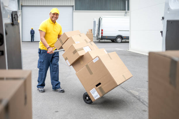 Warehouse worker moving hand truck loaded with cardboard boxes stack, view inside of delivery van. Courier with cart wide shot, freight transportation