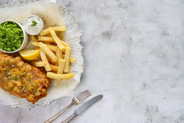 Stock photo showing close-up, elevated view of a white and gold plate with irregular rim lined with greaseproof paper containing a portion of battered cod and chips, with a lemon slice, mushy peas and tartare sauce, against a marble effect background.