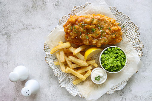 Stock photo showing close-up, elevated view of a white and gold plate with irregular rim lined with greaseproof paper containing a portion of battered cod and chips, with a lemon slice, mushy peas and tartare sauce, against a marble effect background.