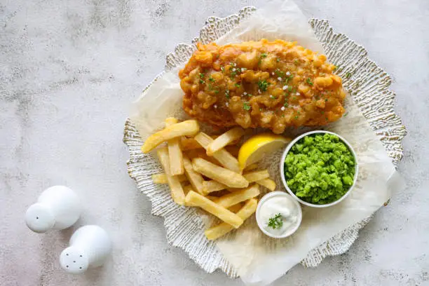 Stock photo showing close-up, elevated view of a white and gold plate with irregular rim lined with greaseproof paper containing a portion of battered cod and chips, with a lemon slice, mushy peas and tartare sauce, against a marble effect background.