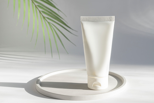 Cosmetic cream tube stands on a ceramic tray under green palm leaf, blank beauty and care product packaging mock-up