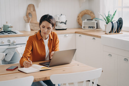 Concentrated young spanish woman is entrepreneur and remote worker. Freelance and distance work concept. Kitchen interior, worktops and cuisines. Daylight through window.