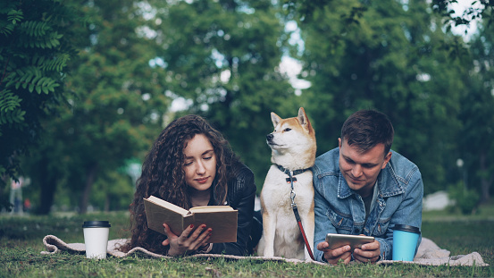 Happy young couple is resting in park, man is using smartphone while woman is reading book, their dog is sitting between them on blanket. People are talking and smiling.