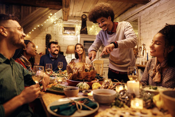 молодой счастливый человек вырезает индейку на день благодарения во время званого обеда с друзьями за обеденным столом. - thanksgiving turkey dinner dinner party стоковые фото и изображения