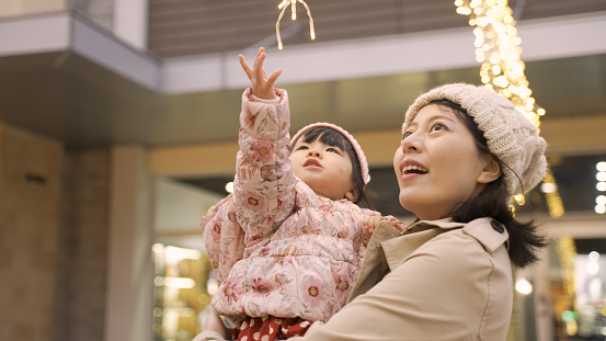 slow motion of cute curious asian toddler baby girl raising up hand trying to touch outdoor Christmas lights in her mother’s arms outside an urban shopping mall