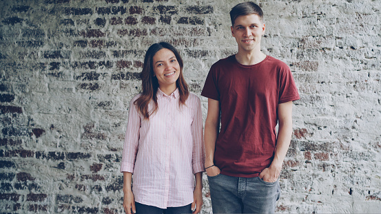 Portrait of attractive couple standing together against brick wall looking at camera and smiling. Happy people, relationship and modern interior concept.