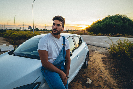 A young man leaning on a car outdoors at sunset