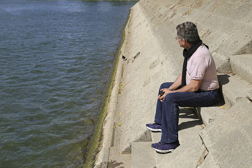 A side view of an adult Brazilian man sitting on stairs near the river in Paris