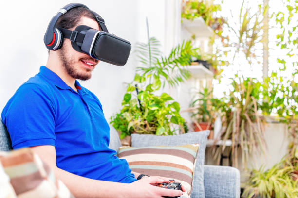 un gars inspiré assis sur un canapé dans le salon porte un casque vr profitez de la réalité virtuelle en ligne jeu cyber space world simulation. - three dimensional recreational pursuit behavior ecstatic photos et images de collection