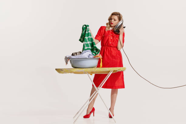 服でいっぱいのボウルを持って立ち、電話のように鉄の上で話す美しい女性のポートレート - 1950s style iron stereotypical housewife women ストックフォトと画像