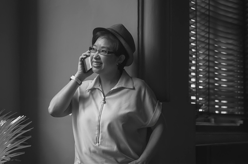 Smiling Asian adult woman in vintage style talking on smartphone while leaning against the wall inside of living room in black and white style