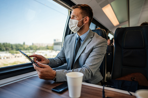 Handsome middle-aged businessman using his tablet computer while traveling with high-speed train. Modern and fast travel concept. He is wearing protective face mask as protection during virus pandemic.
