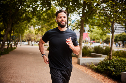 Smiling young man in sportswear listening to music on wireless earbuds while running along a tree lined footpath in an urban park
