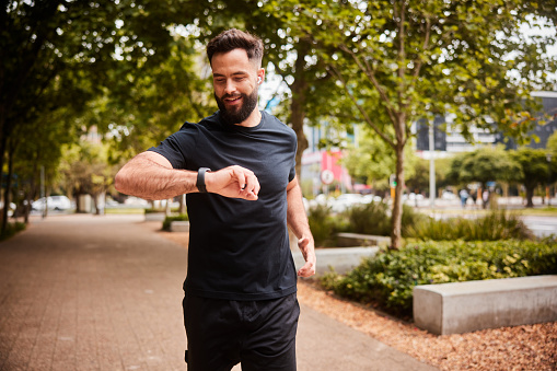 Smiling young man in sportswear and wearing earbuds checking his fitness tracker while out for a run along a path in a city park