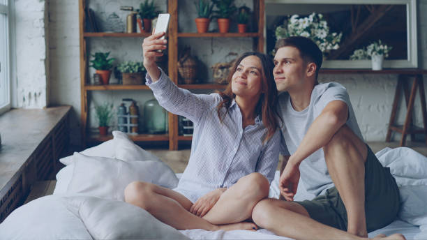istock Cheerful loving couple is taking selfie with smartphone looking at camera, posing and making funny faces while sitting together on bed at home. Technology and relationship concept. 1436975784