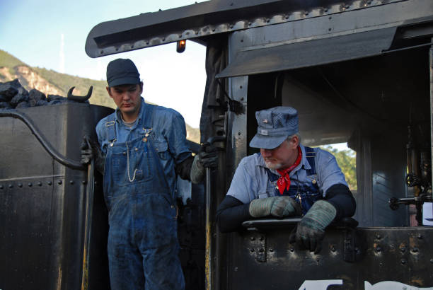 A portrait of a rugged looking middle-aged Caucasian male construction worker with a gray beard, wearing an aluminum hard hat and has a leather tool belt over his shoulder, looking off into the distance, with an old rusted steel plate and corrugated steel background. He is also wearing a blue denim shirt with a off white Henley underneath.
