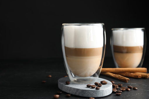 Delicious latte macchiato, cinnamon sticks and coffee beans on black table, space for text