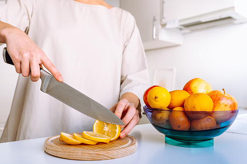 woman's hands cut lemon slices on cutting wooden board to make refreshing lemon drink, bowl of fresh fruits stands nearby
