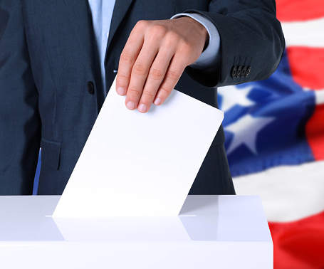 Election in USA. Man putting his vote into ballot box and American flag on background, closeup